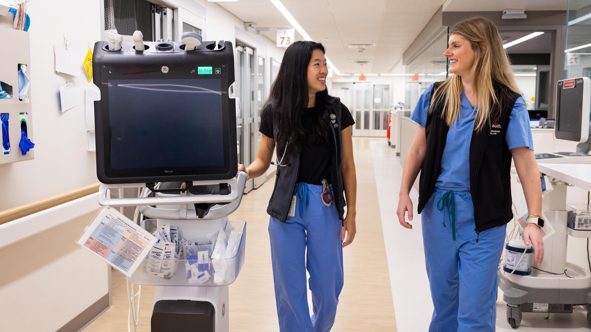 Two medical providers talk while walking through a hall and guiding a cart. 