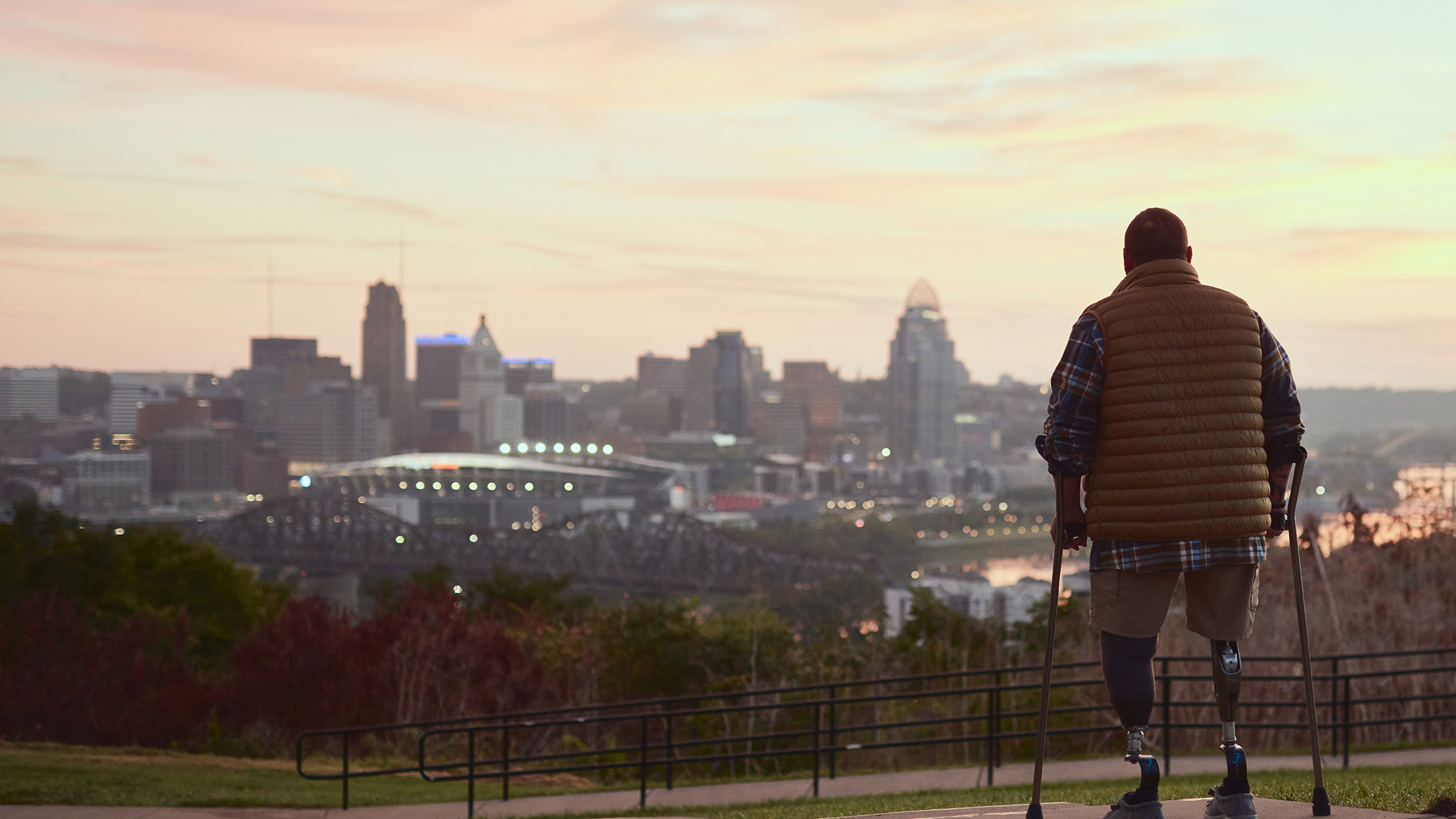 Man with prosthetic legs overlooks downtown Cincinnati from a hill at dusk.