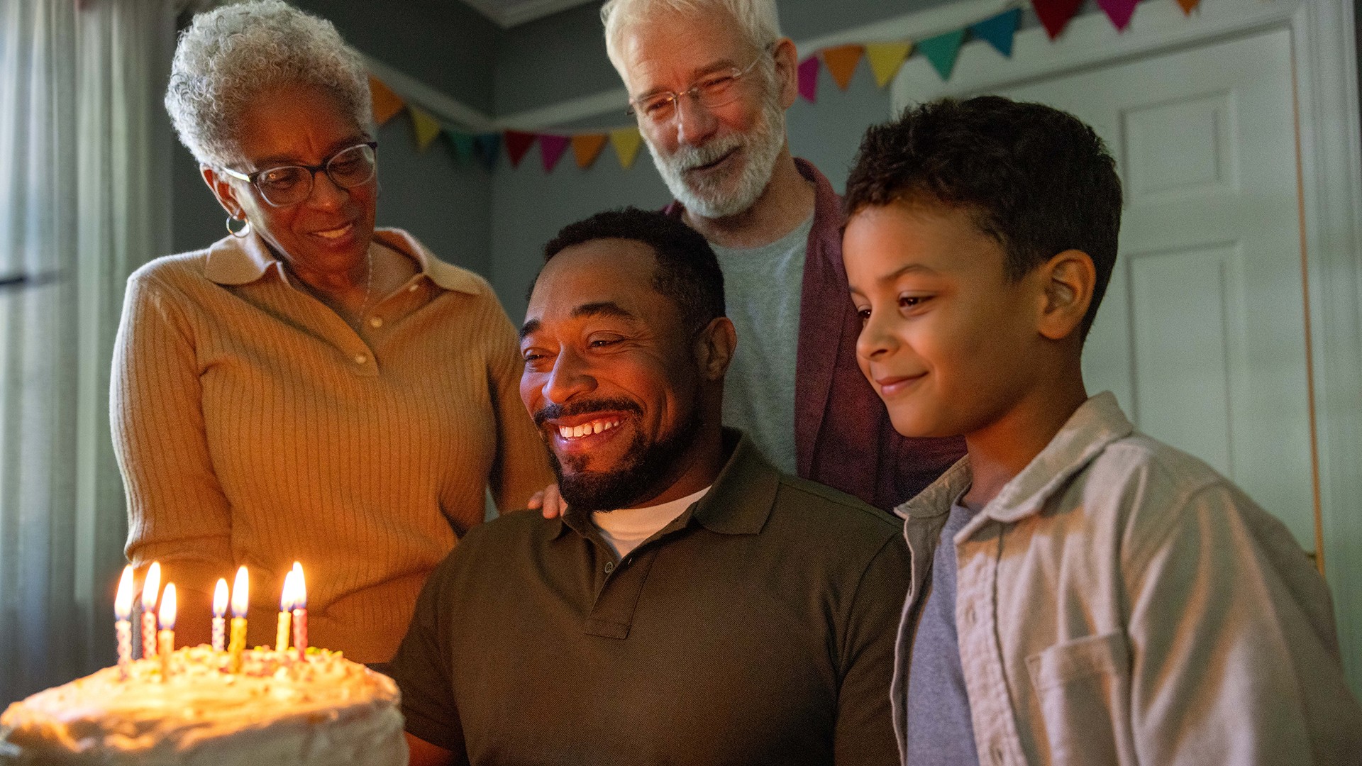 Man stares at a birthday cake with candles with a big smile and his family around him.