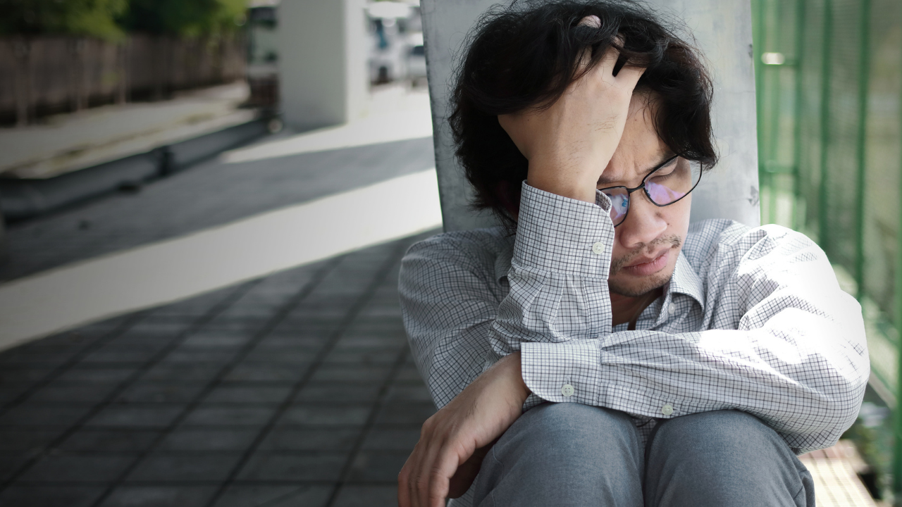 Man sits on the ground outside holding his head seemingly experiencing distress