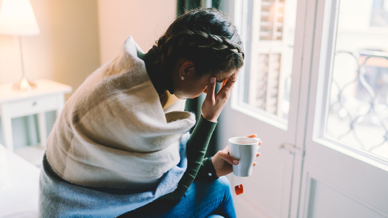 woman sitting on a bed with a cup of tea head holds her head in discomfort