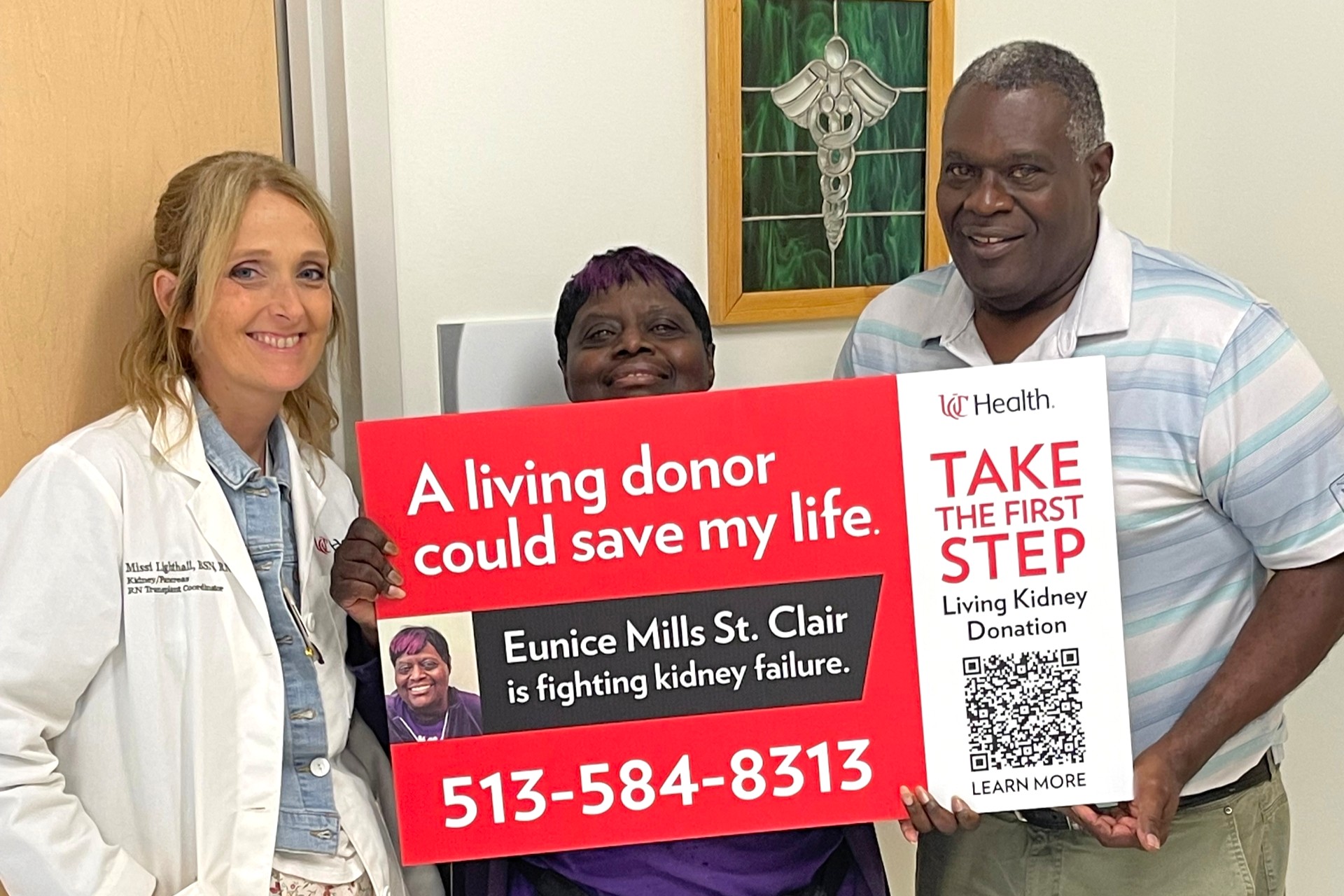 A physician stands next to a patient holding a sign with a family member to her side. 