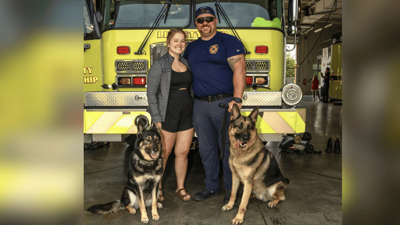 Austin, his fiance, and his two dogs stand together in front of a firetruck at the fire station.