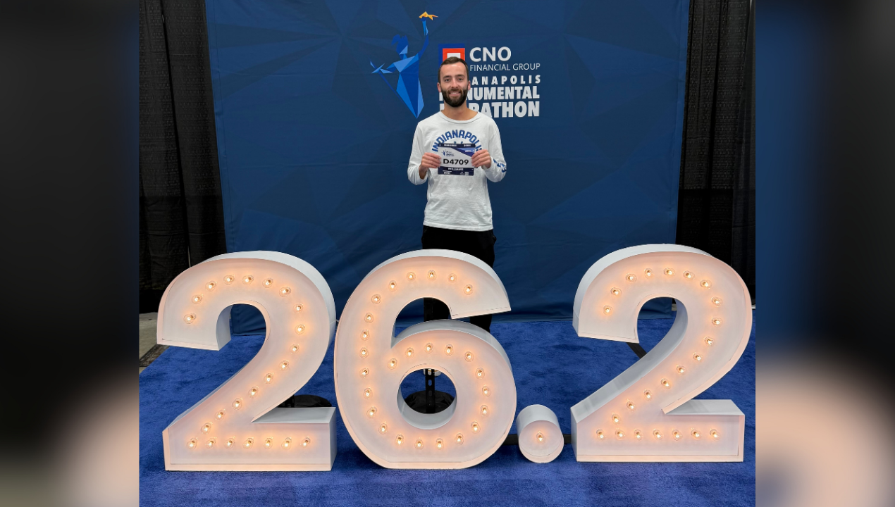Daniel shows his running bib in front of a 26.2 sign while preparing to run his next marathon