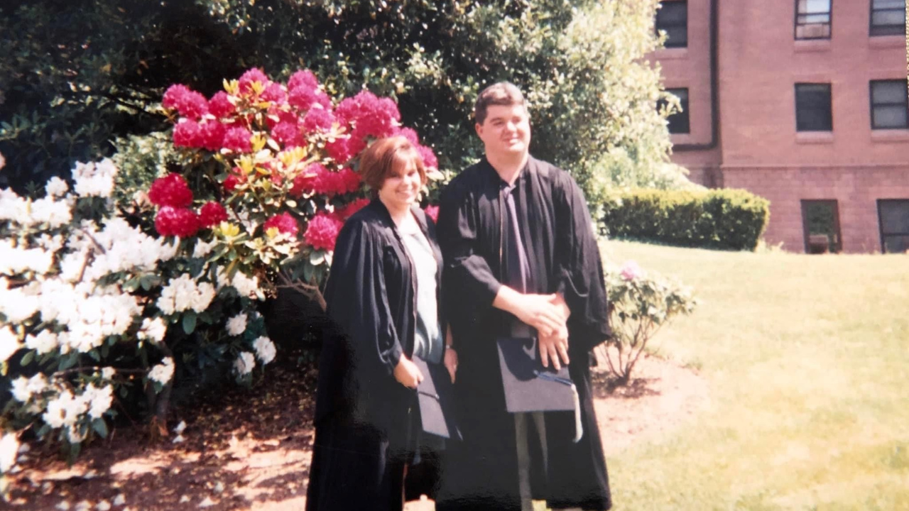 Hilary and Amy Debelak dressed in graduation gowns and standing in front of flowers.