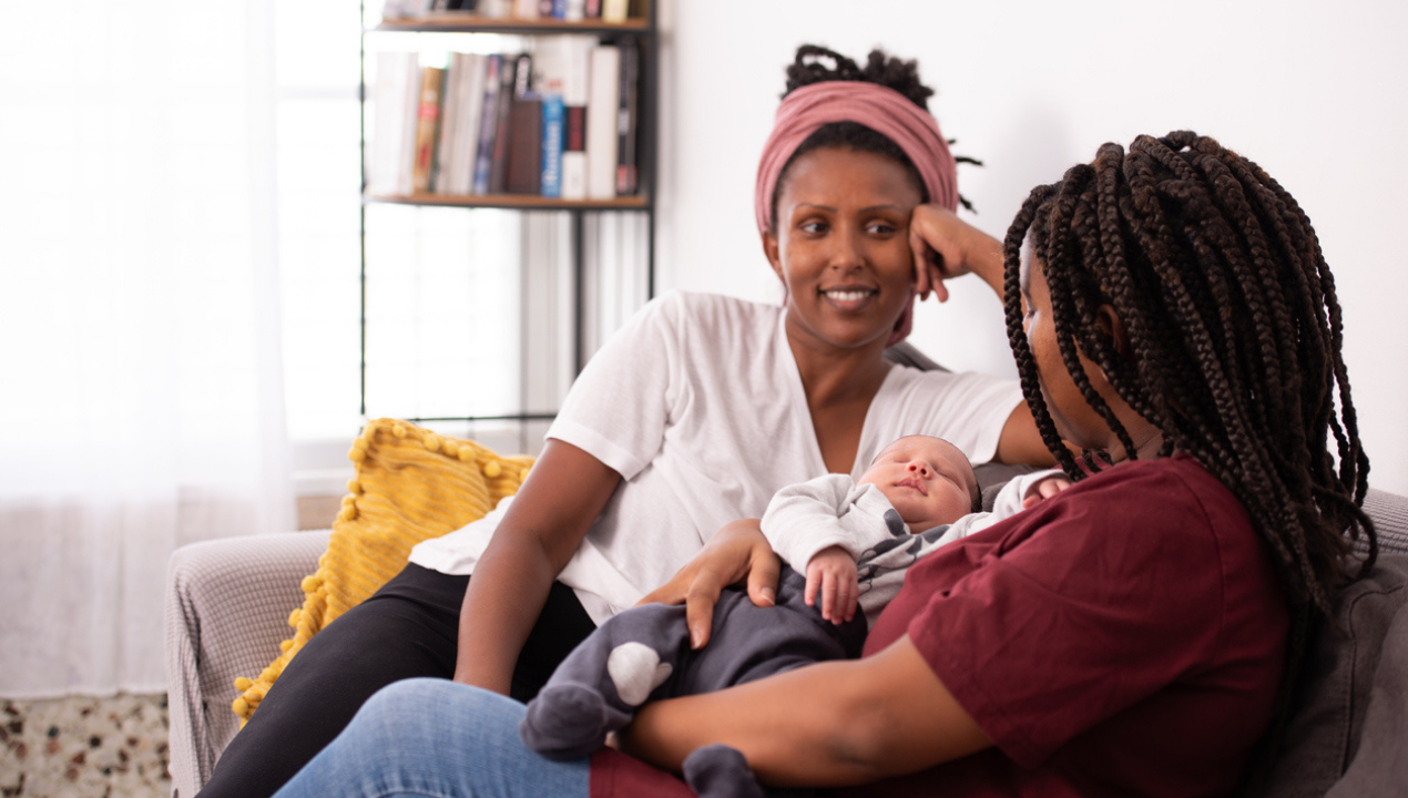 Two women sit on couch with one woman holding a new baby