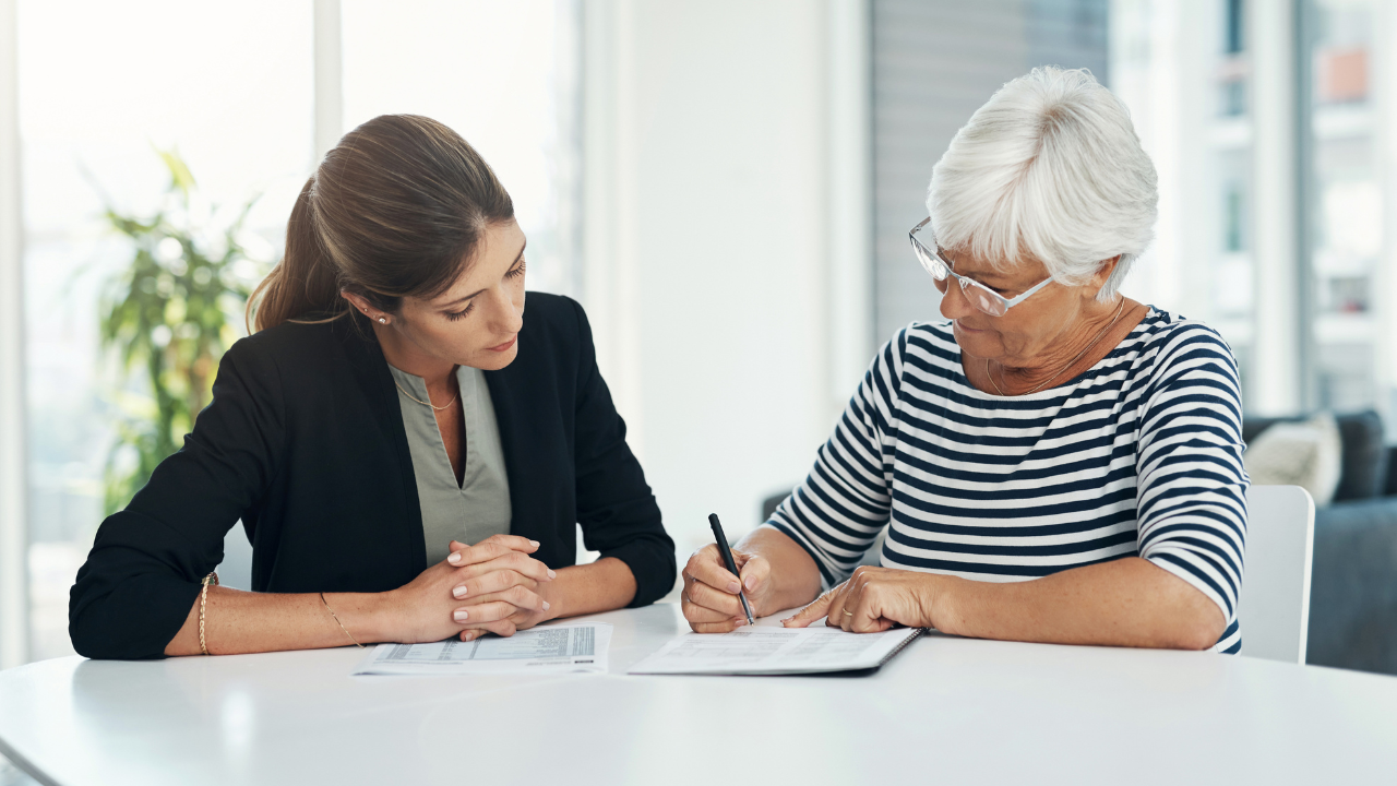 Two women sit at a table and one signs paperwork while the other looks on. 
