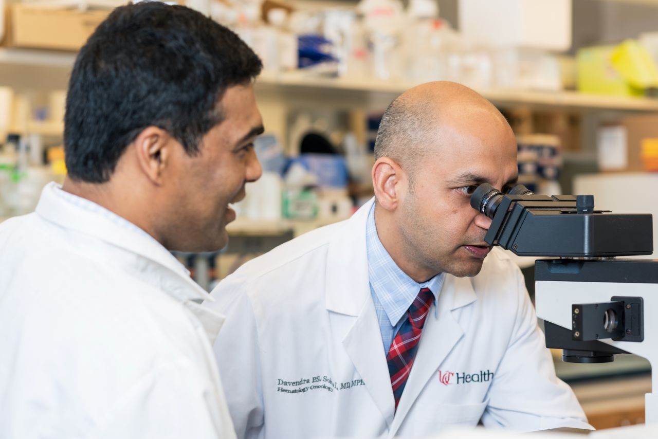 Physician looks through a microscope while another physician looks on.