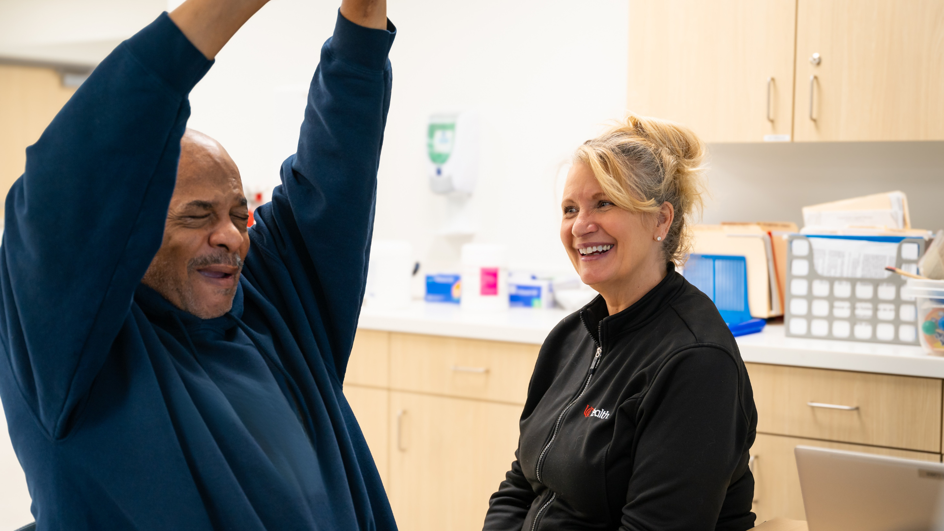 Therapist smiles at a patient as he lifts his arms in the air and smiles. 