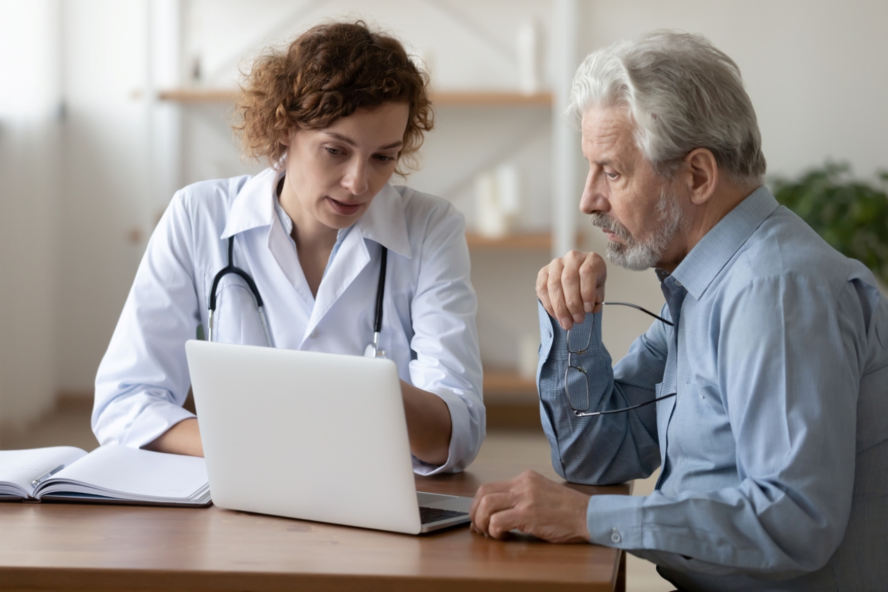 Physician and patient sit at a desk and talk while looking at a laptop. 