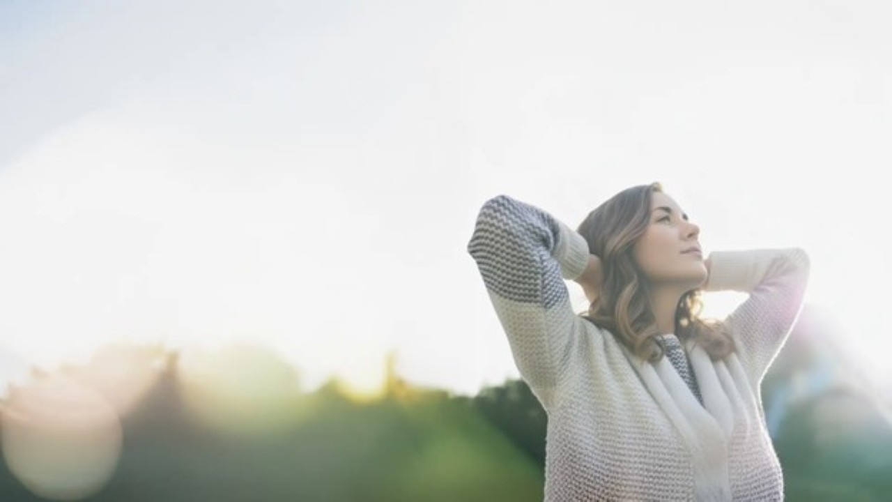 Woman stands outside in the sun and smiles as she stretches her arms above her.