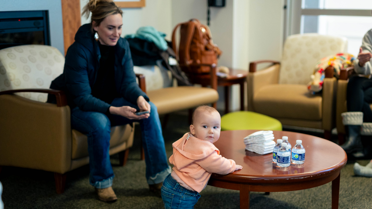 Mom watches as her baby stands at a coffee table during a Baby Cafe gathering