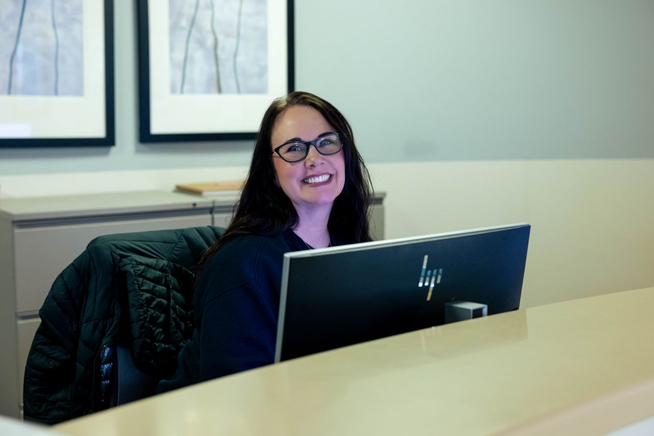Medical provider sits next to a desk with a computer monitor in front of her and smiles at the camera.