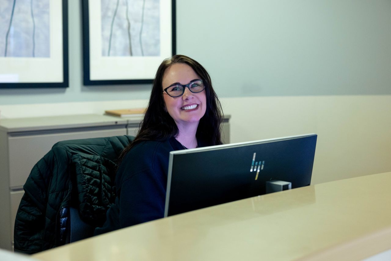 Medical provider sits next to a desk with a computer monitor in front of her and smiles at the camera.